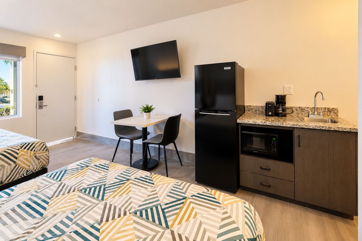 Een kleine hotelkamer met een bed, een eettafeltje met stoelen, een zwarte koelkast, een magnetron en een koffiezetapparaat.