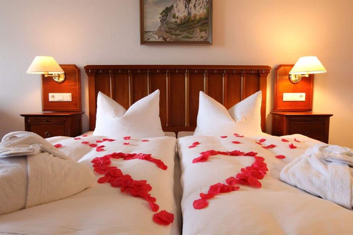 Quarto de hotel com duas camas, cada uma com roupa de cama branca e pétalas de rosa vermelha dispostas em formato de coração. Possui cabeceiras e mesas de cabeceira de madeira, com luminárias.