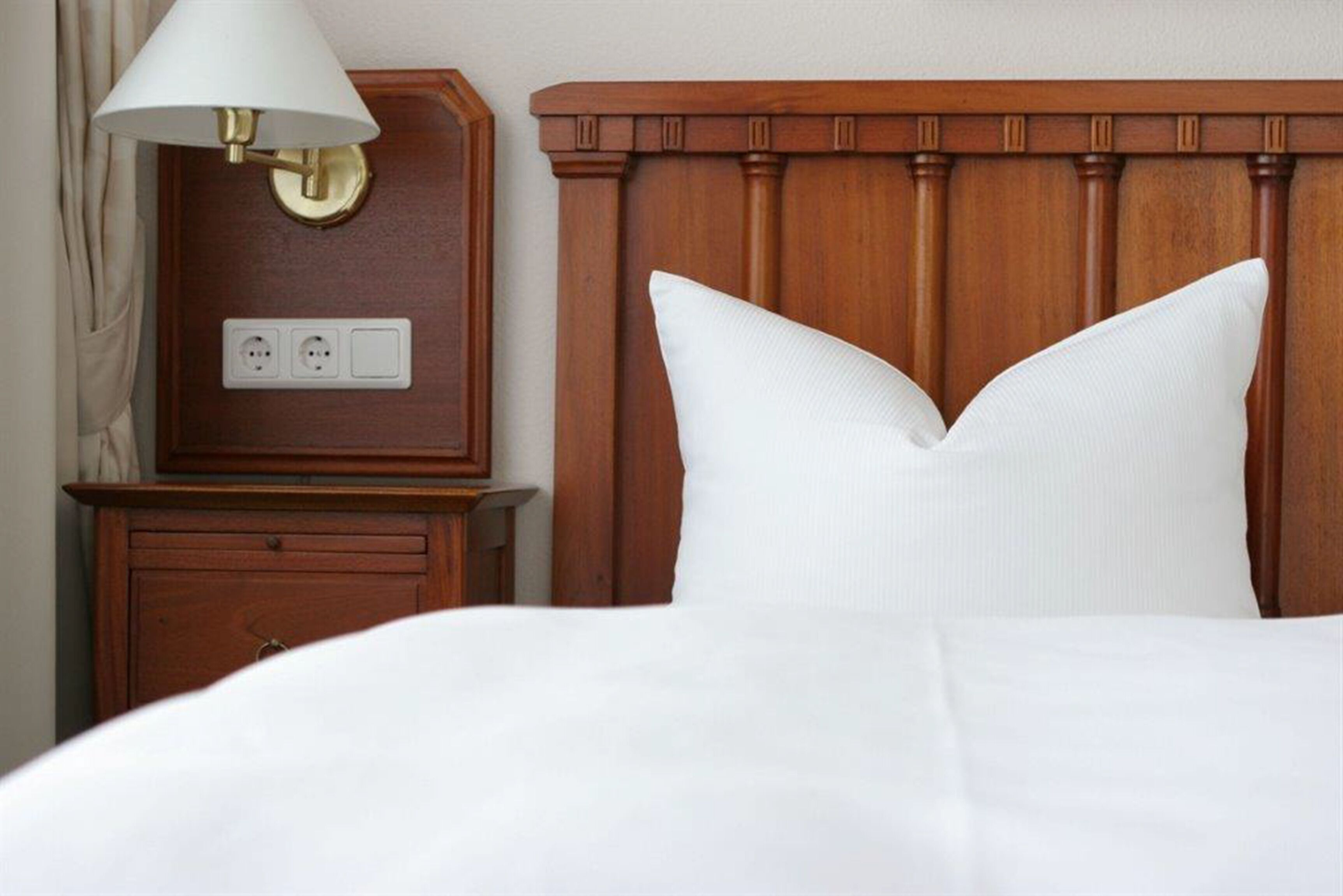 Un letto rifatto con lenzuola bianche, una testiera in legno, un comodino con una lampada e una presa di corrente.
