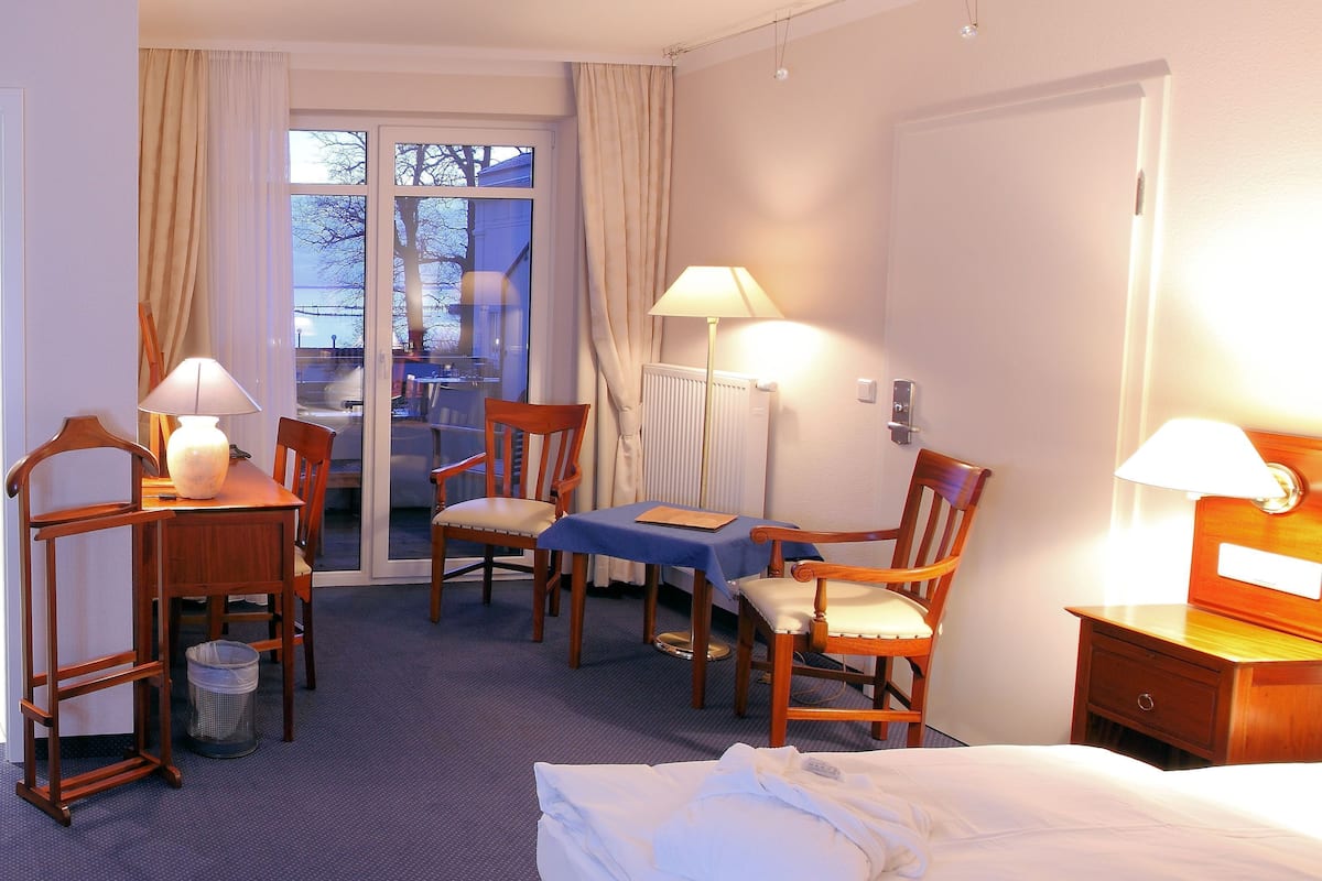 Quarto de hotel com cama, escrivaninha com abajur, cadeira, mesa com abajur e vista para o exterior pela janela.