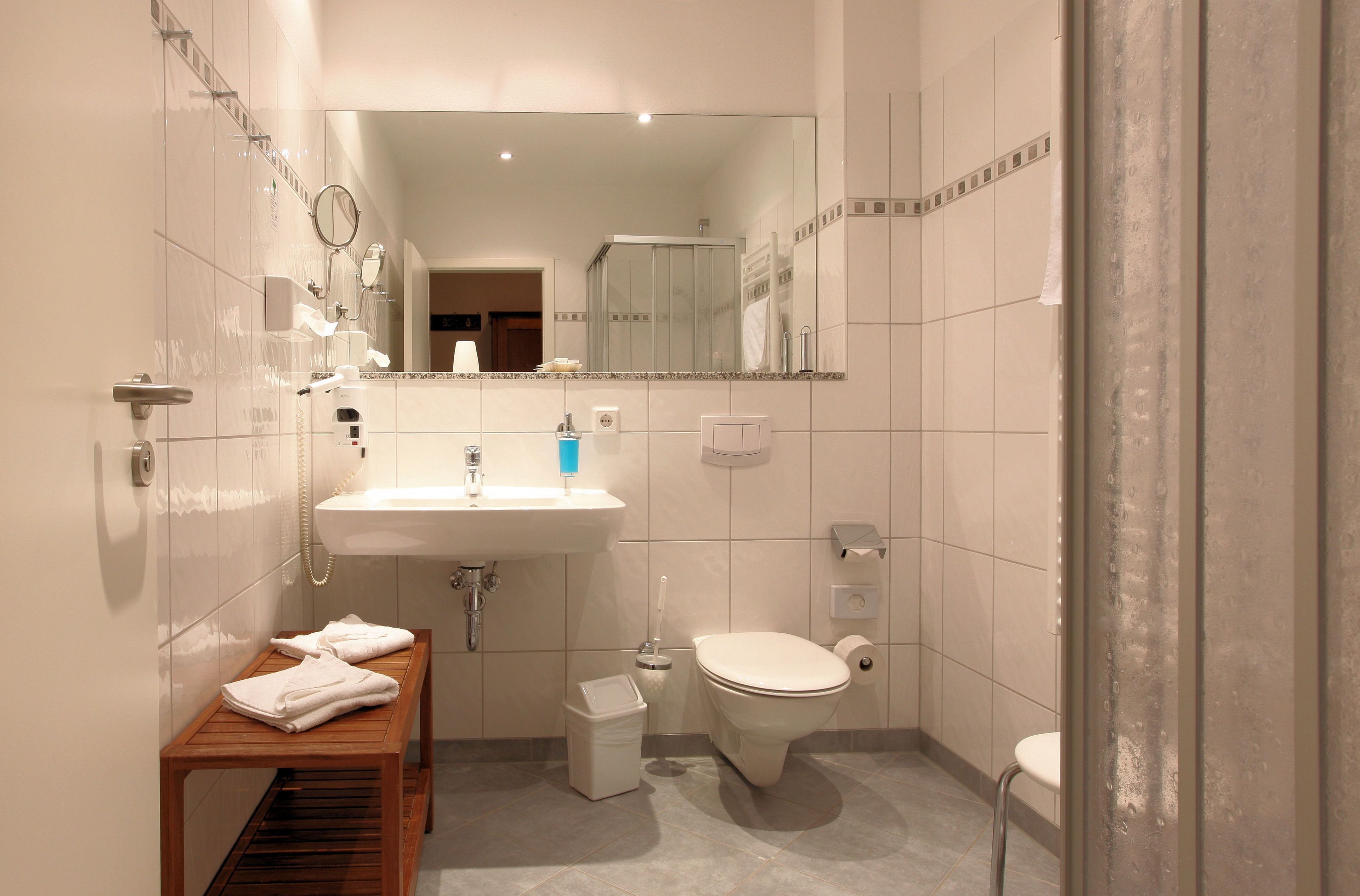 A modern bathroom with a sink, toilet, and shower. There is a bench with towels and a trash bin.