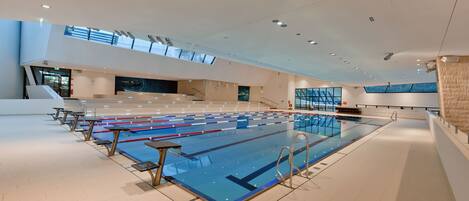 A large indoor swimming pool with multiple lanes, a spectator area, and a modern architectural design with large windows and a high ceiling.
