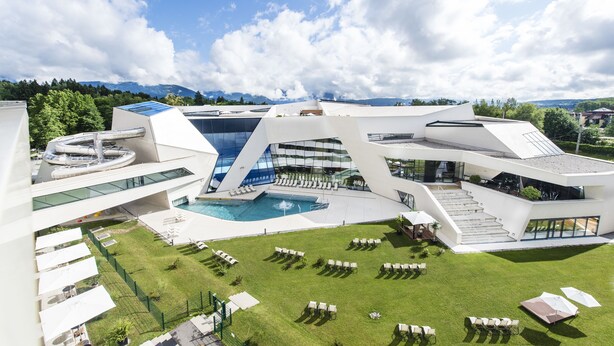 A modern building with a pool, outdoor seating, and a view of mountains. - Thermenhotel Karawankenhof (Villach)