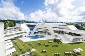 A modern building with a pool, outdoor seating, and a view of mountains. - Thermenhotel Karawankenhof (Villach)