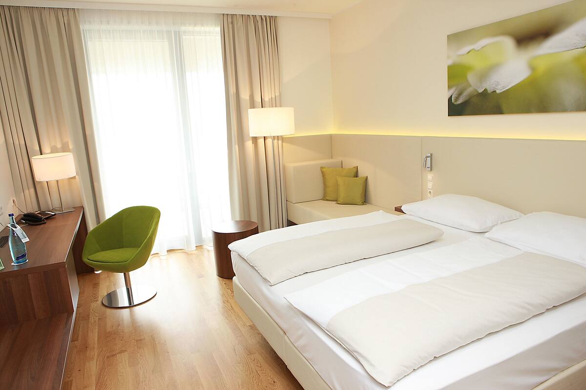 Una habitación de hotel con una cama, un escritorio, una silla y un ventanal con cortinas.