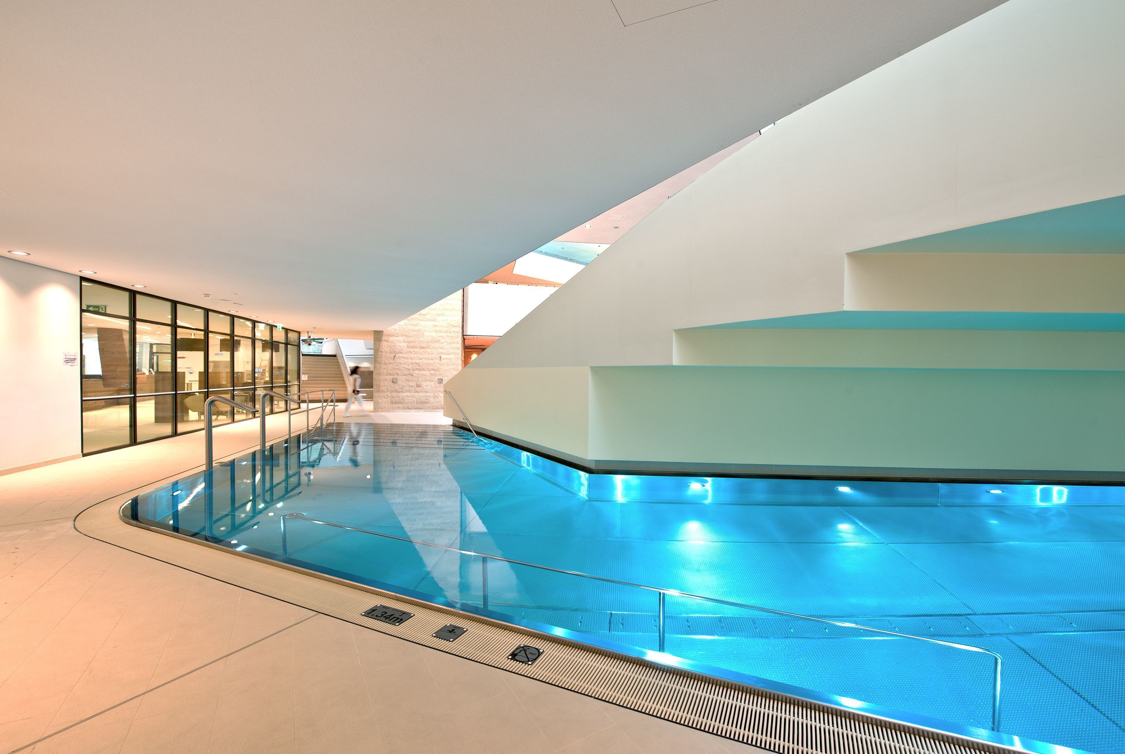 Una piscina interior con paredes de vidrio transparente, piso de baldosas y un diseño arquitectónico moderno con techo inclinado y una escalera que lleva a un nivel superior.
