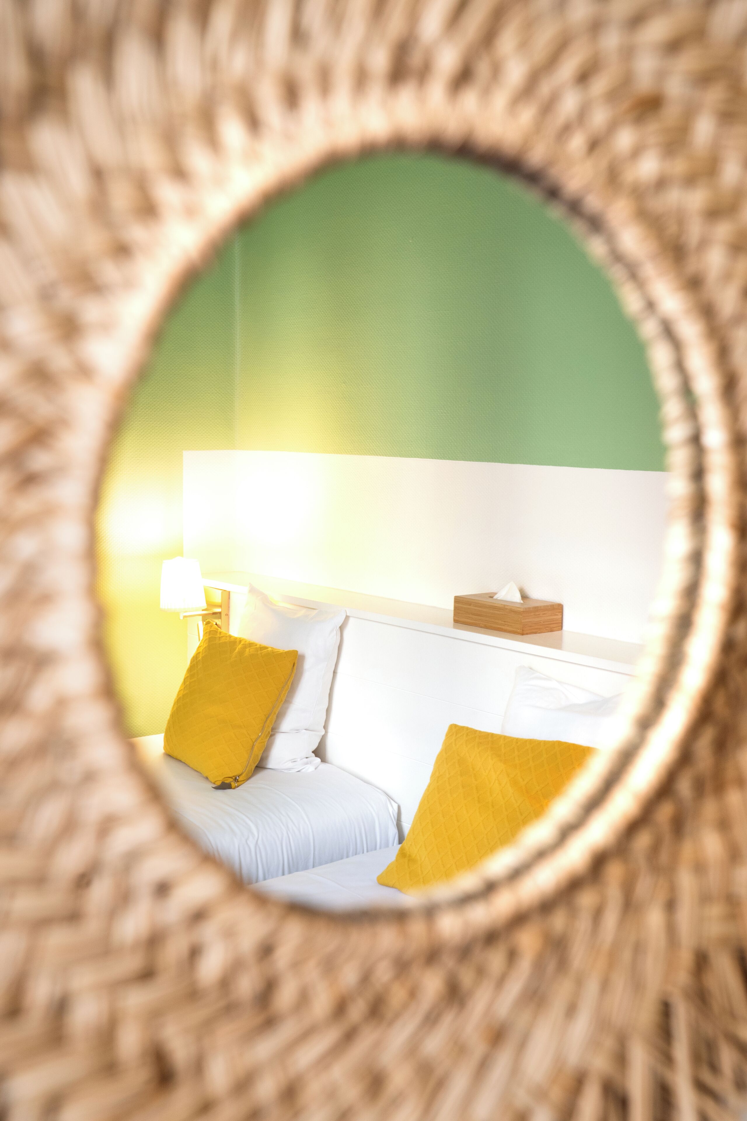 Un espejo circular refleja una cama con ropa de cama blanca y dos almohadas amarillas, una lámpara de noche y un caja de madera sobre una pared verde.