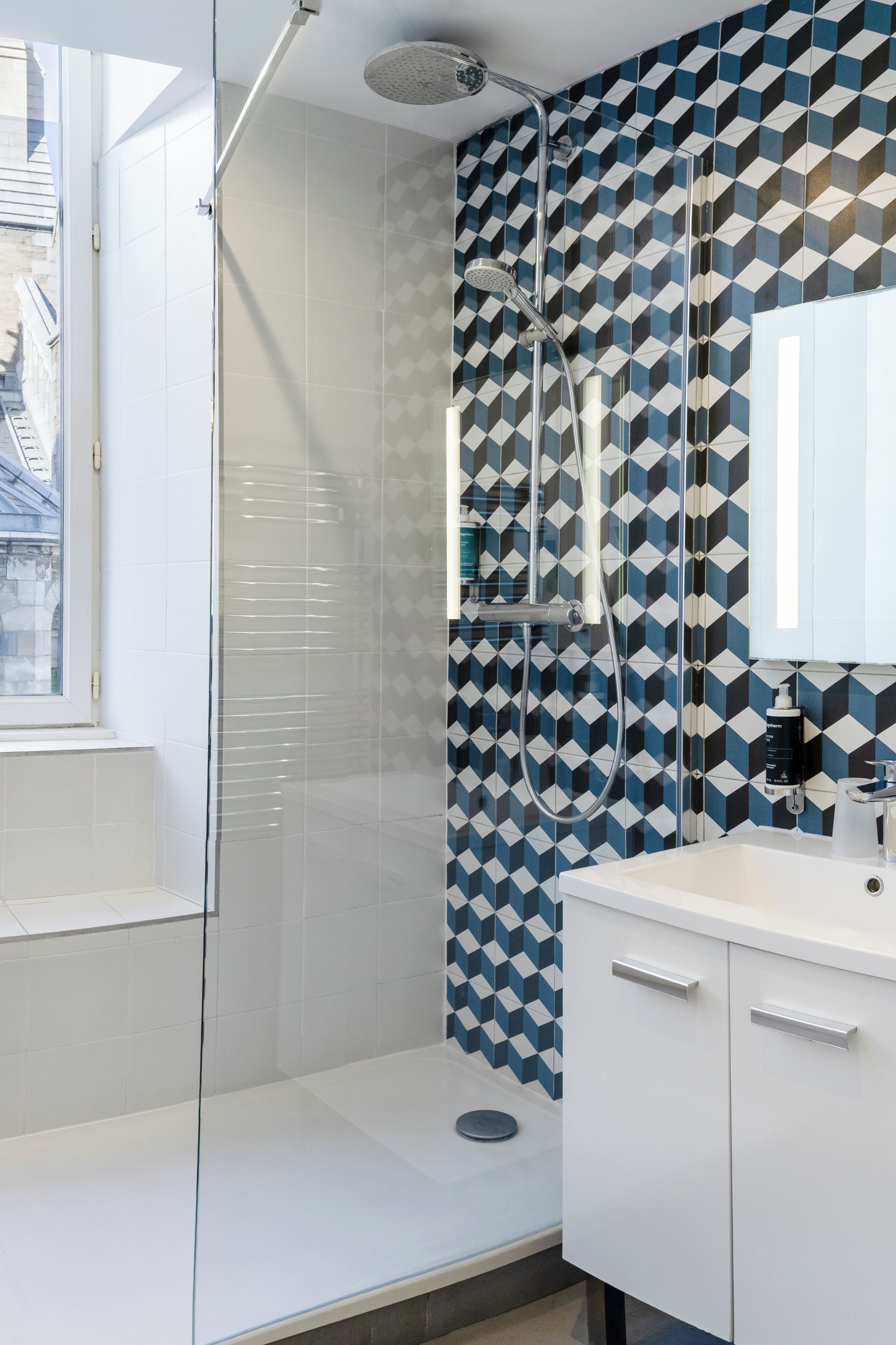 A modern bathroom with a geometric tile backsplash, a glass-enclosed shower, and a white vanity with a mirror.