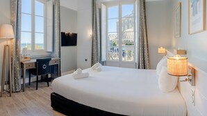A hotel room with a large bed, a desk, a chair, and a TV. There are curtains on the windows, and a lamp is on the bedside table. - Hôtel de L'Océan et Restaurant - Coeur de Ville (Biarritz)