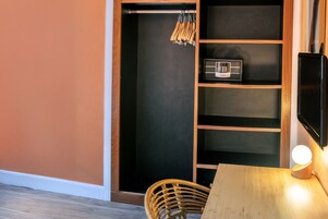 Una habitación con una mesa de madera, una silla de mimbre, un estante negro con una radio y un armario de madera con perchero.
