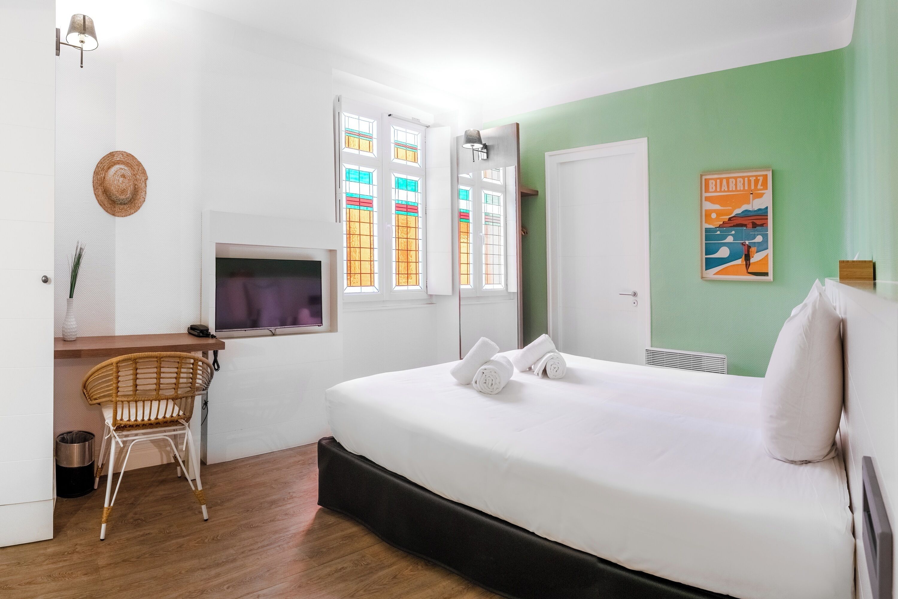 Un dormitorio con cama, escritorio, silla, televisor y ventana con vidrios de colores.