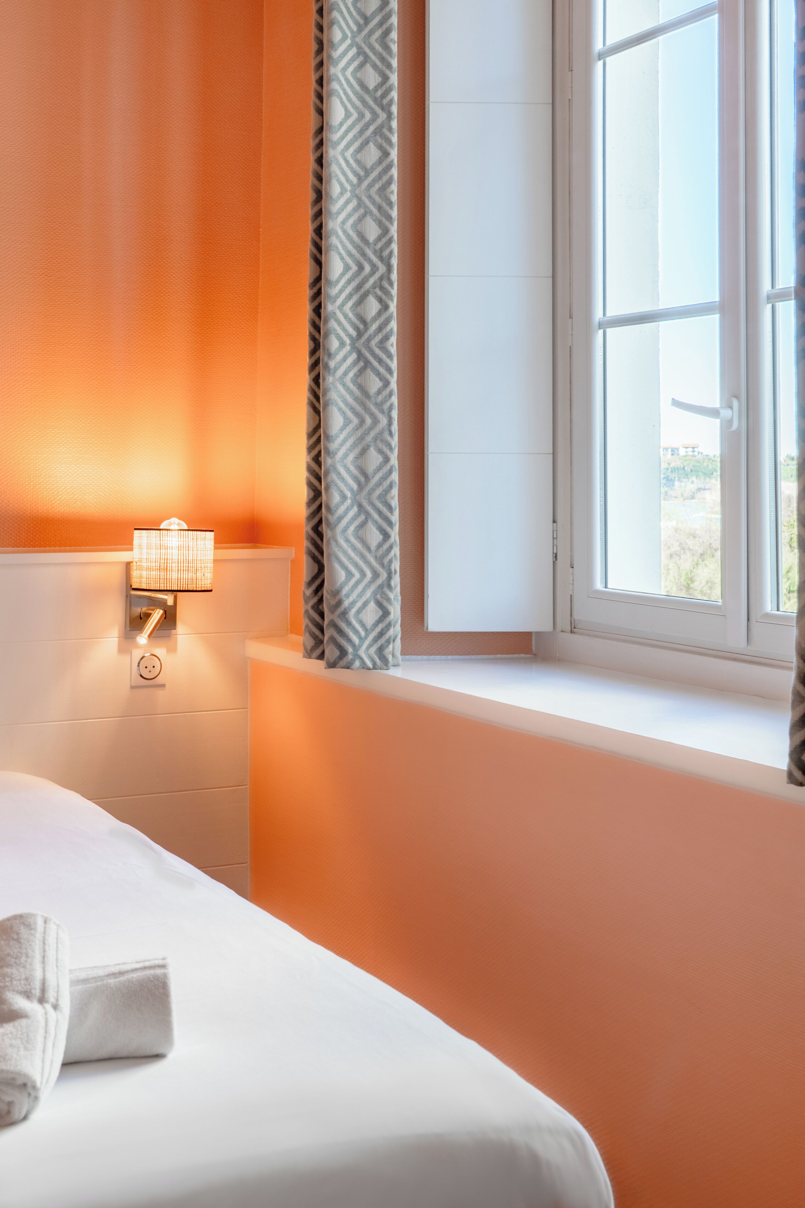 Een hotelkamer met een bed, nachtkastje, lamp, raam met gordijnen en uitzicht op de omgeving.