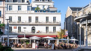 A hotel with outdoor seating, a sign reading "HOTEL DE L'OCEAN," and a classical building nearby. - Hôtel de L'Océan et Restaurant - Coeur de Ville (Biarritz)