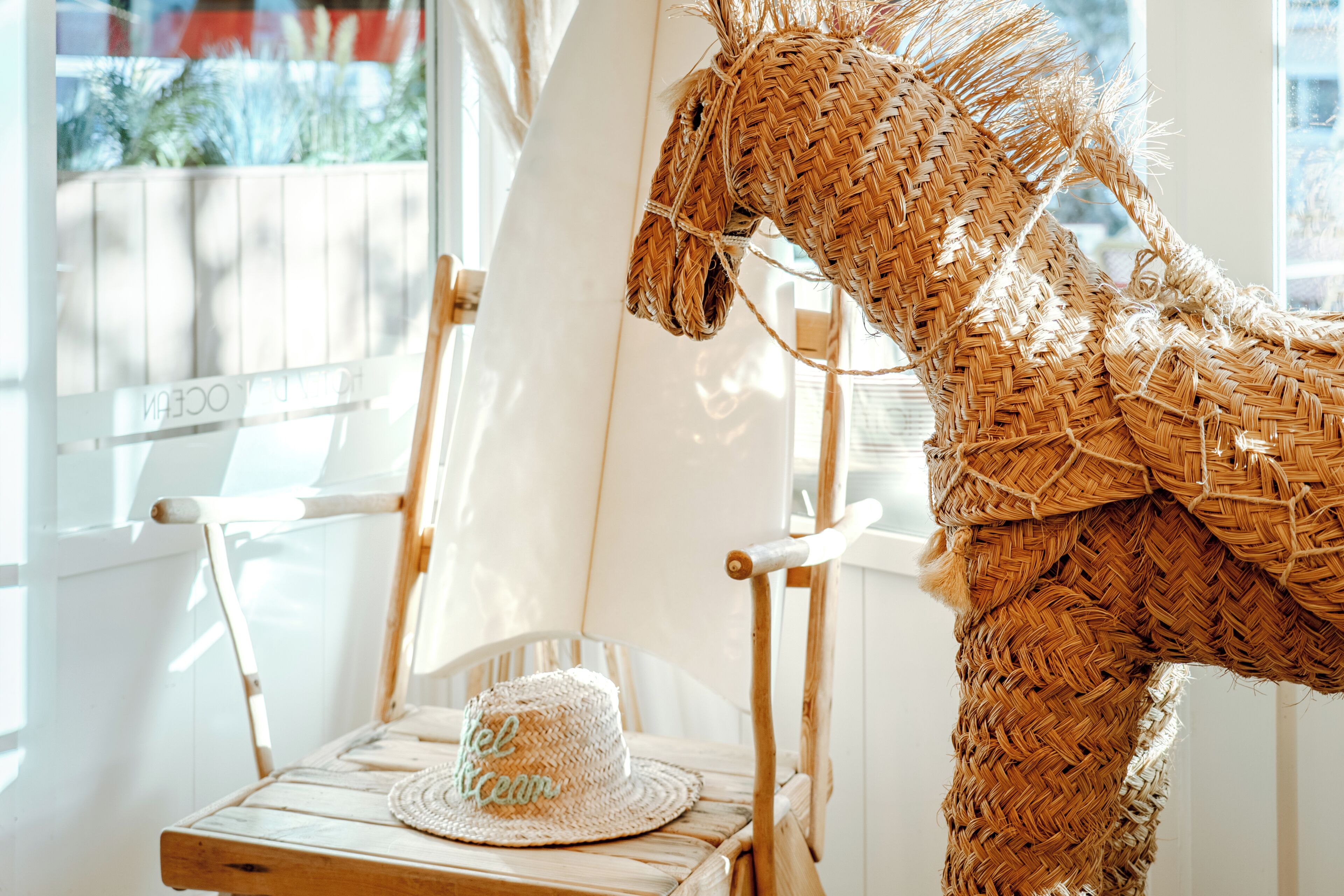Una escultura de caballo de mimbre, un sombrero de mimbre y una silla de madera en una habitación con una ventana blanca y otra silla blanca.