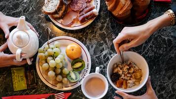 A breakfast setting with a plate of bread, meats, and fruits, a teapot, and a bowl of cereal on a marble table.