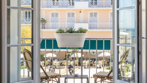 A hotel room with a large arched window, a bed, and a view of a courtyard with outdoor seating and a building with balconies. - Hôtel de L'Océan et Restaurant - Coeur de Ville (Biarritz)