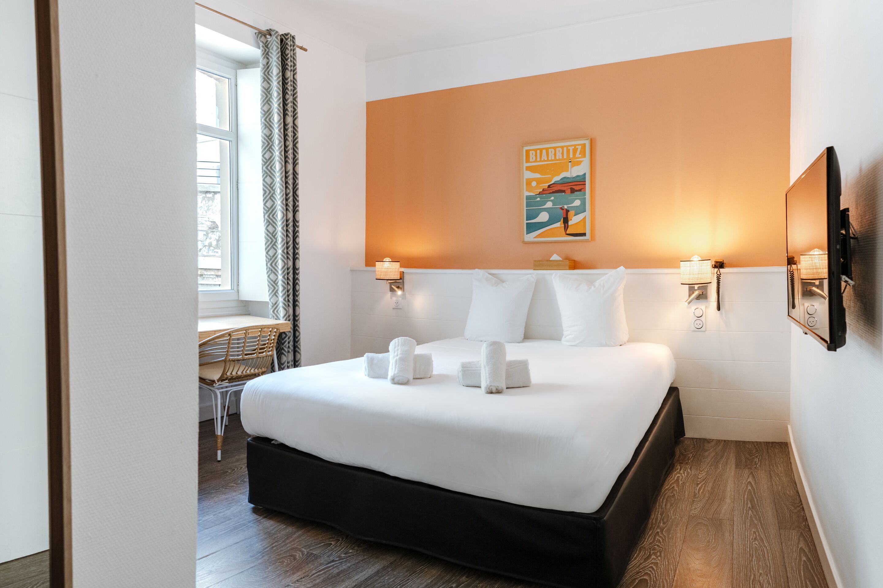 A hotel room with a large bed, two bedside lamps, a painting on the wall, and a window with curtains.