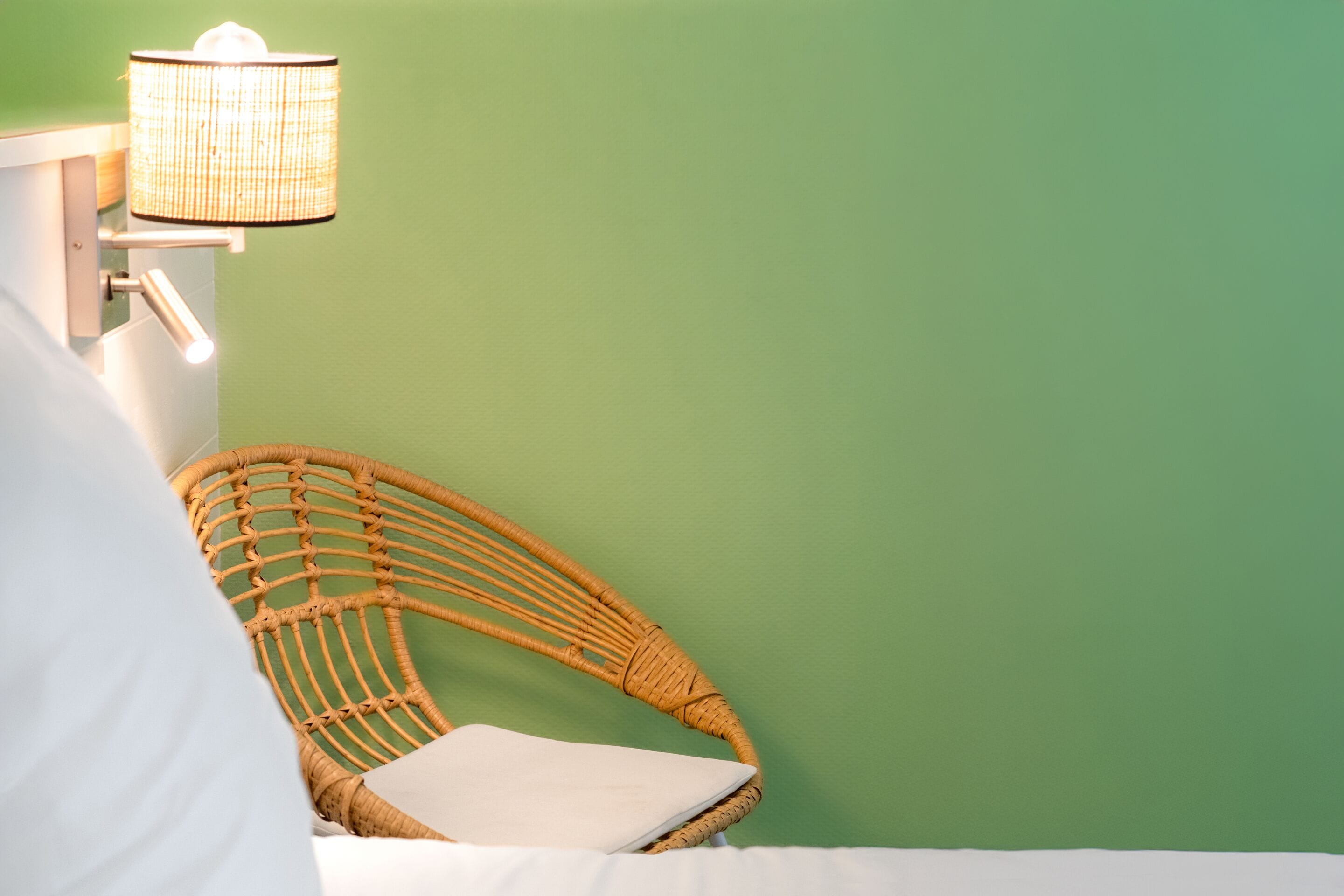 A wicker chair with a white cushion against a green wall, a white pillow on a bed, and a wall-mounted lamp.