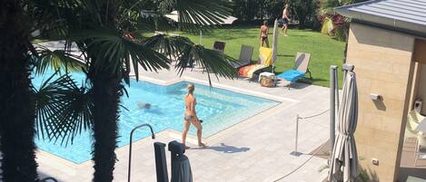 Una piscina con solárium de baldosas, rodeada de palmeras y sillones de descanso.