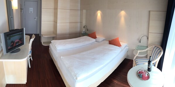 A hotel room with a bed, a desk with a computer, a chair, a small table with a bottle and glasses, and a door in the background.
