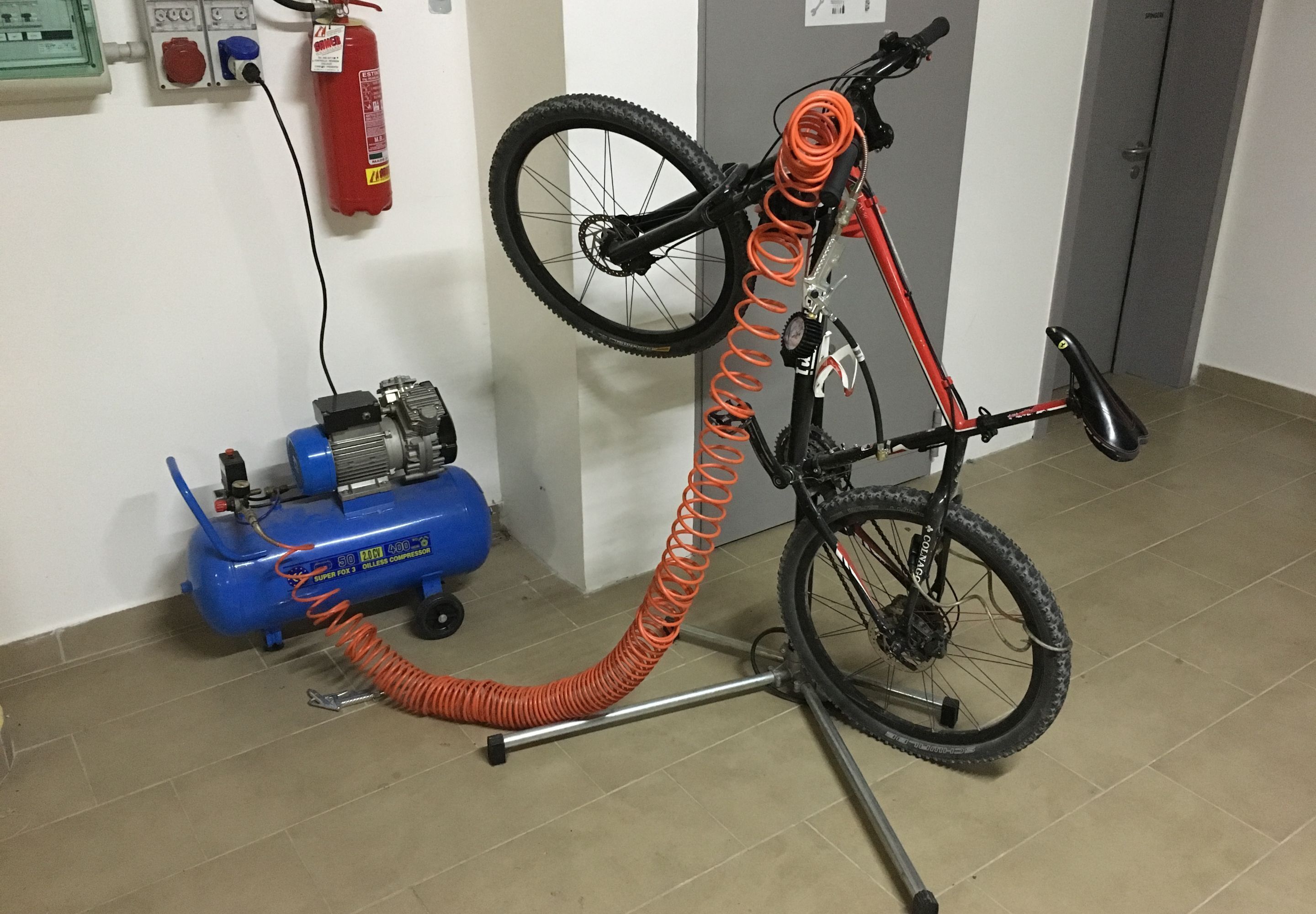 A bicycle mounted on a stand with an orange coiled hose, a blue compressor, and a fire extinguisher on the wall.