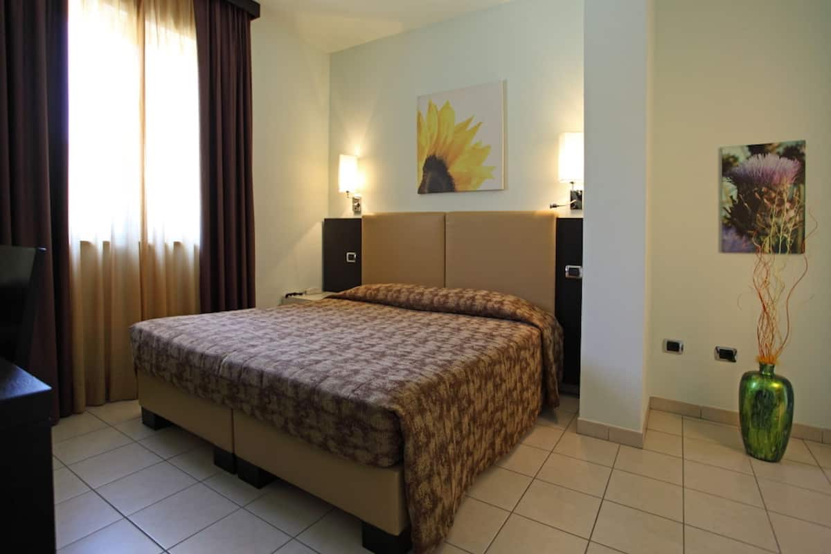 A hotel room with a large bed, two bedside tables, a nightstand, a TV, a window with curtains, and artwork on the walls.
