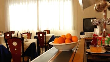 Une salle à manger d’hôtel avec des tables dressées pour les repas, un saladier rempli d’oranges sur le comptoir et un téléviseur allumé au fond.