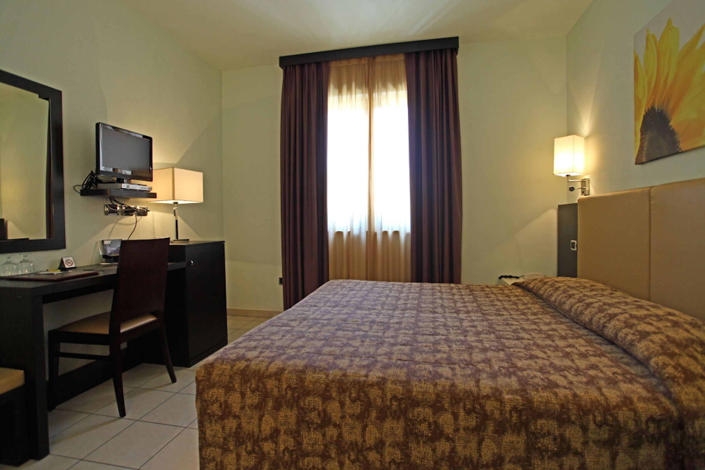 Une chambre d’hôtel comprenant un lit, un bureau avec une chaise, une télévision et une fenêtre avec des rideaux.