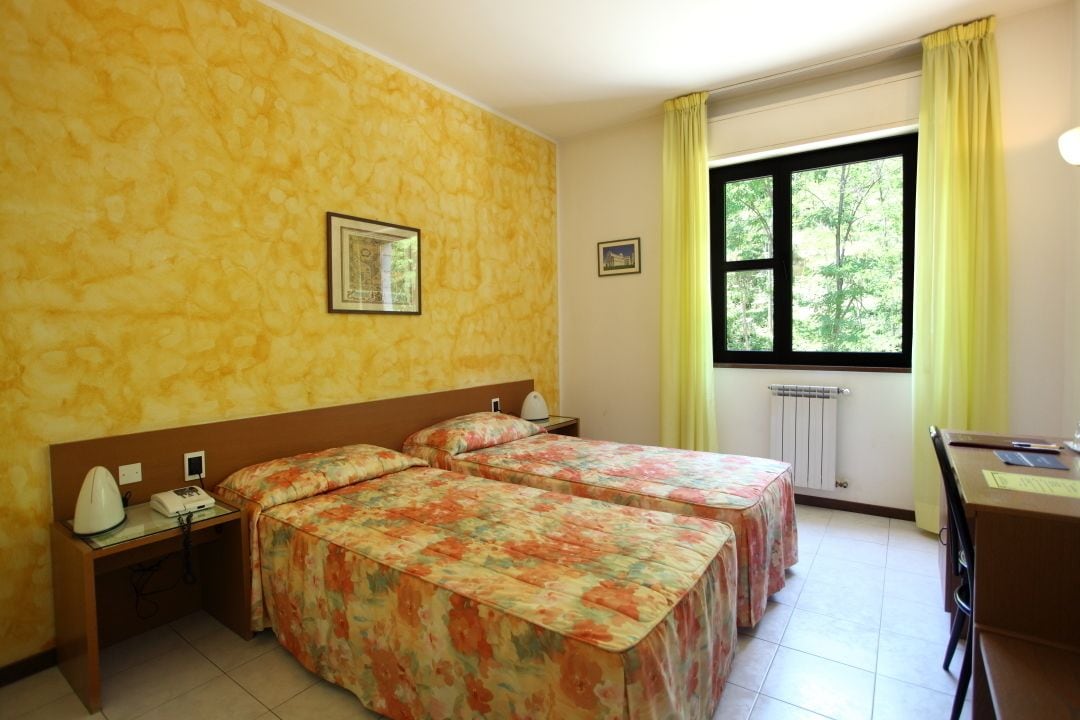 A hotel room with two beds, a desk, a chair, a radiator, and a window with curtains.