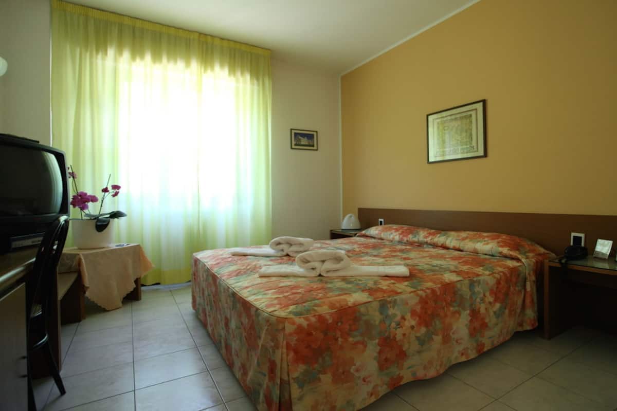 A hotel room with a bed, a desk, a chair, a television, a nightstand, and a window with curtains.