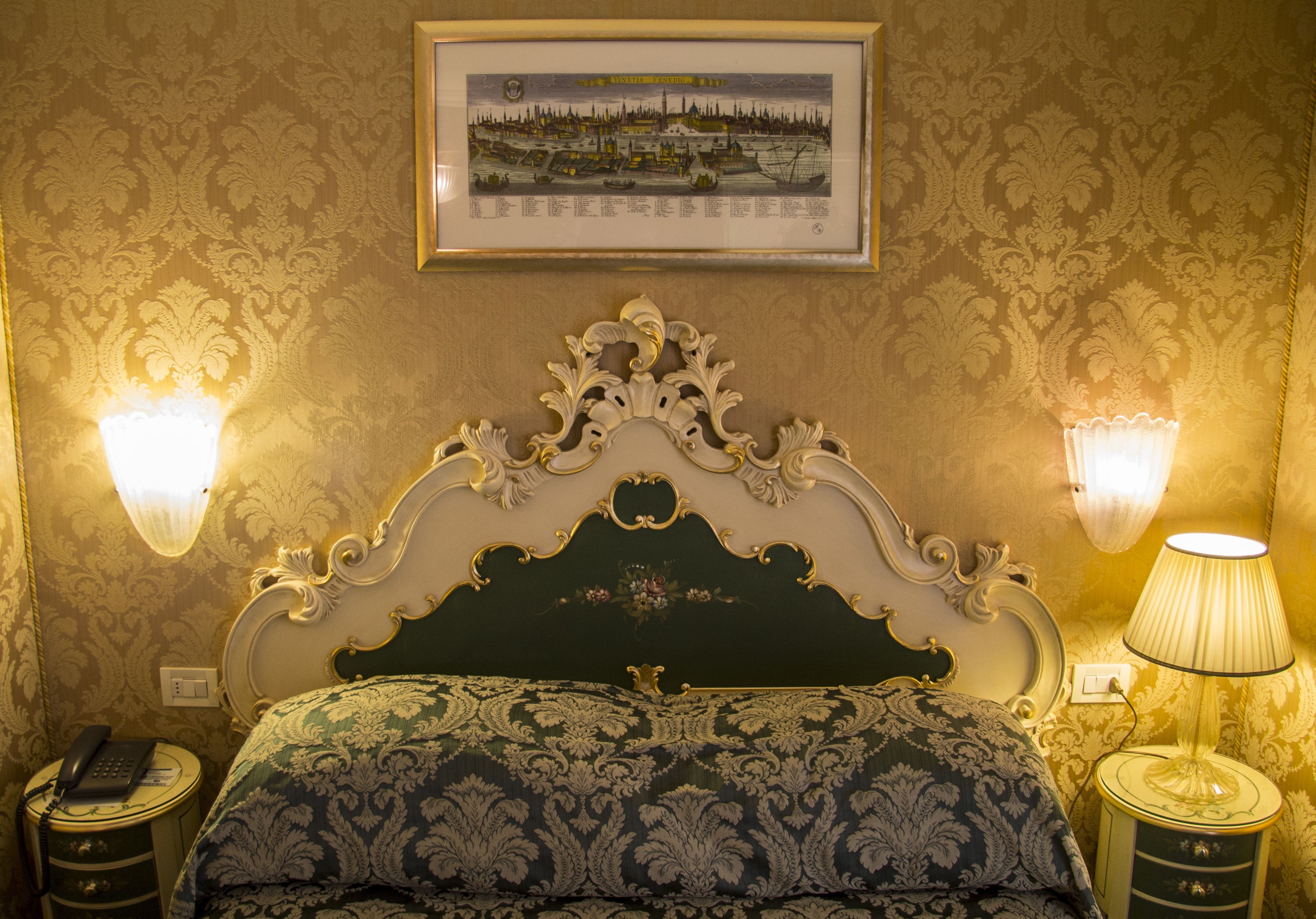 Une chambre avec un lit recouvert d’un tissu vert, un sommier décoratif, deux lampes fixées au mur et un tableau encadré accroché au mur.