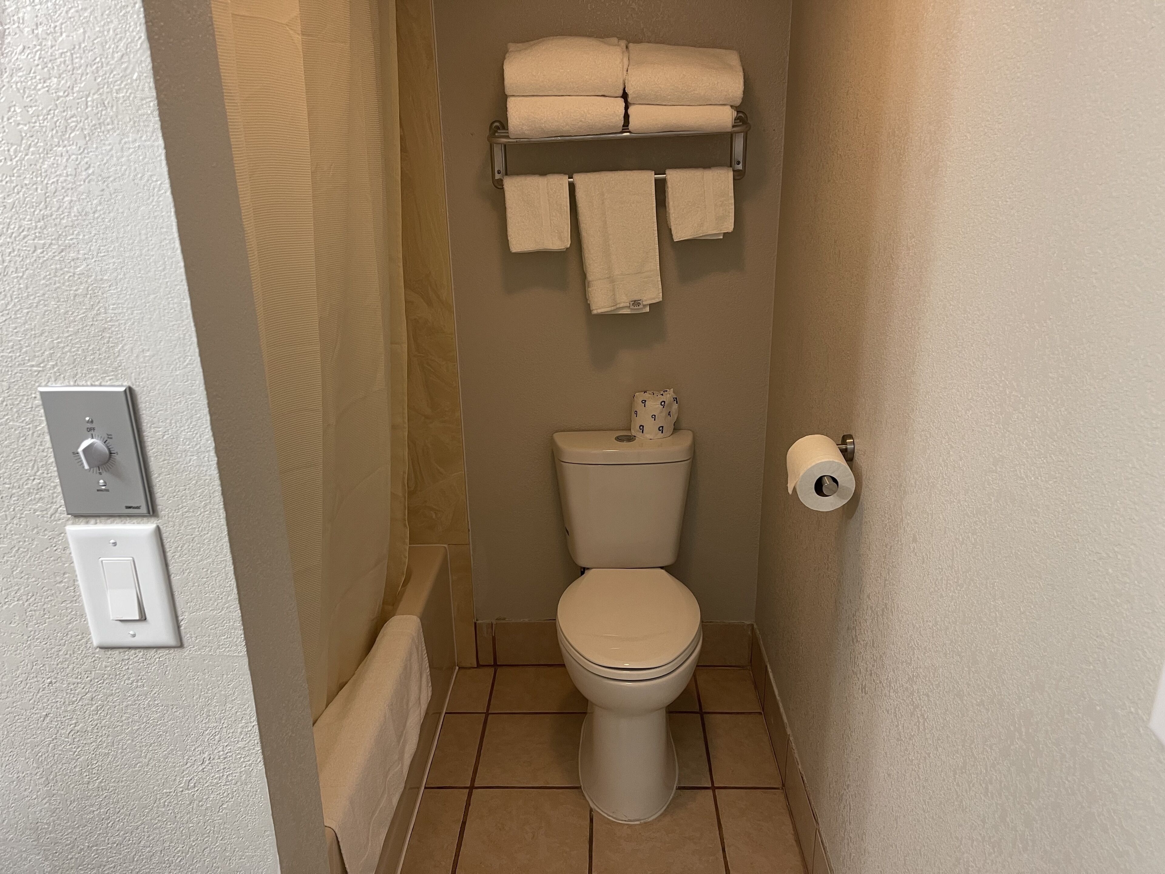 Ein kleines Badezimmer mit Toilette, Handtuchhalter mit gefalteten Handtüchern, Toilettenpapierhalter und Lichtschalter.