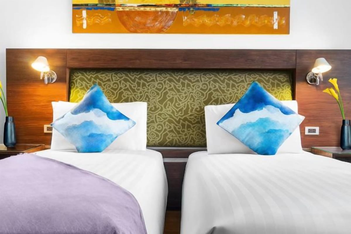 A hotel room with two beds, a wooden headboard, patterned wallpaper, and wall-mounted lamps.