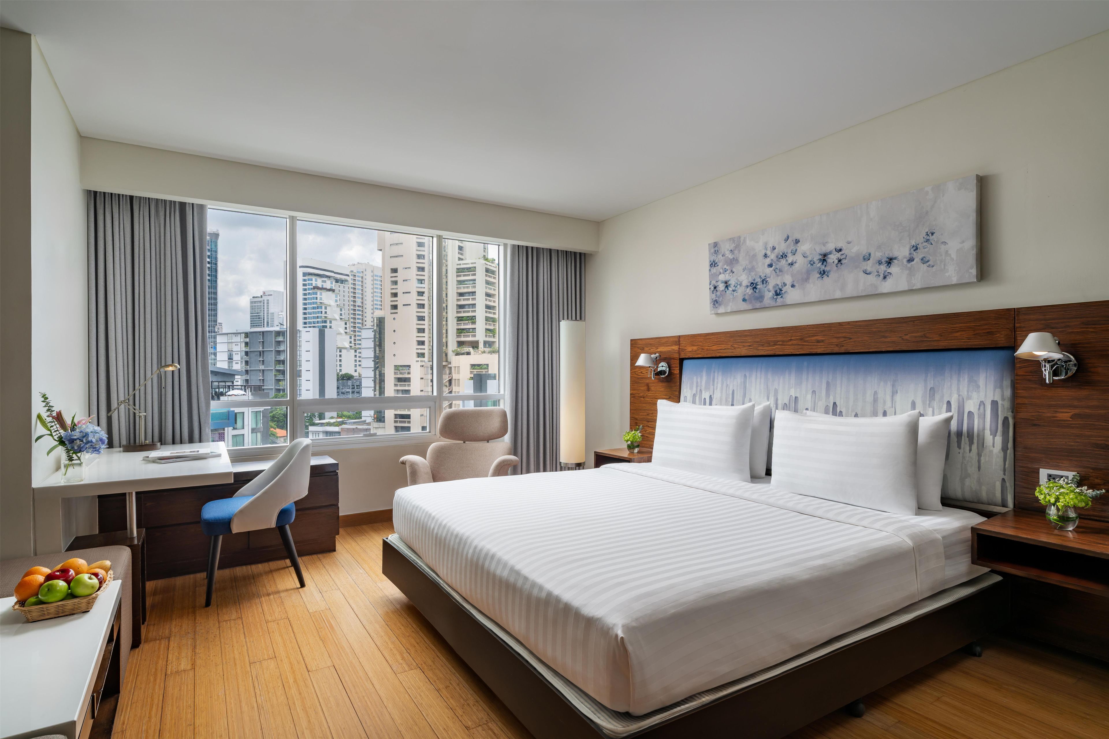 Habitación de hotel con una cama grande, un escritorio, una silla y vistas de la ciudad a través de las ventanas.
