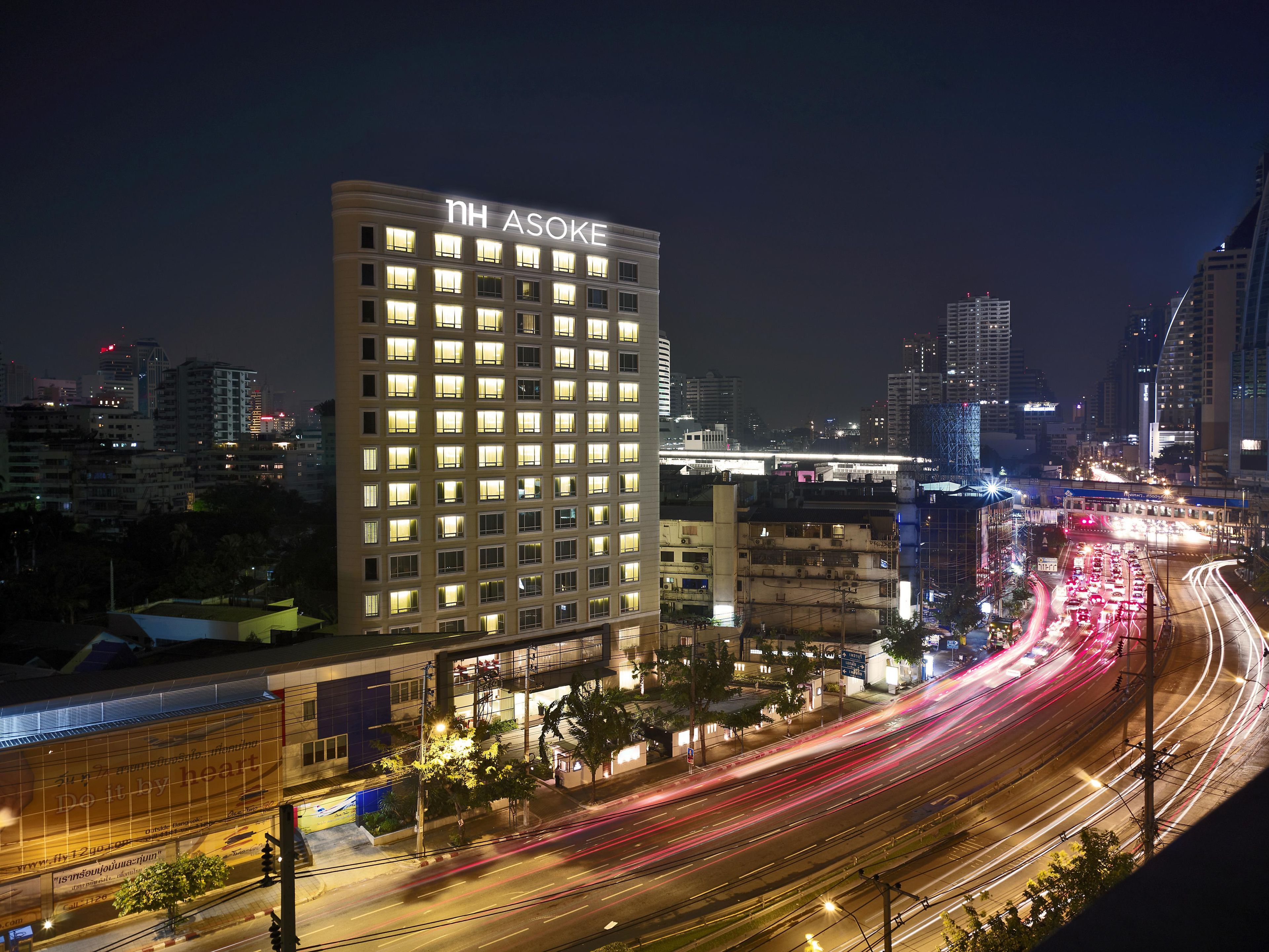 Un immeuble de grande hauteur dont les fenêtres sont illuminées et sur lequel on peut lire, en haut, le nom « NH ASOKE ».