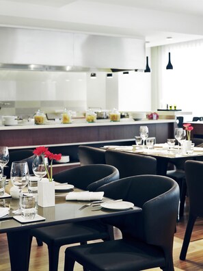 A modern dining area with black leather chairs, white table settings, and a kitchen in the background. - NH Bangkok Asoke (Bangkok)