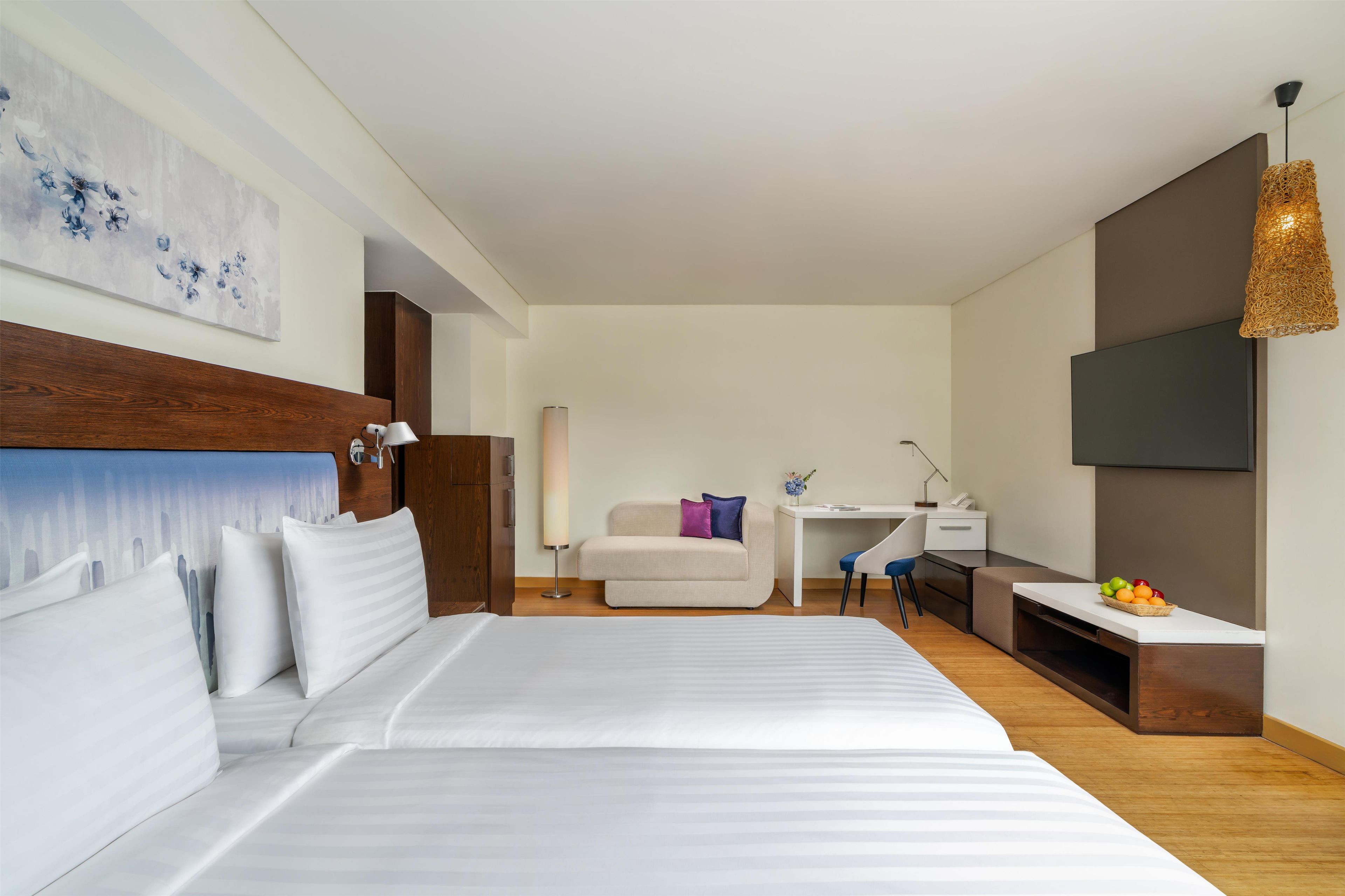 A modern hotel room with a large bed, a desk, a chair, a TV, and a fruit basket.