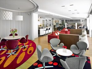 Eine moderne Hotelhalle mit Barbereich, Sitzgelegenheiten und einer im Hintergrund sichtbaren Küche.