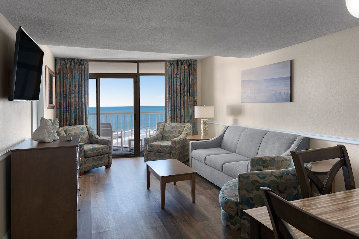A hotel room with a balcony, a sofa, a coffee table, and a TV.
