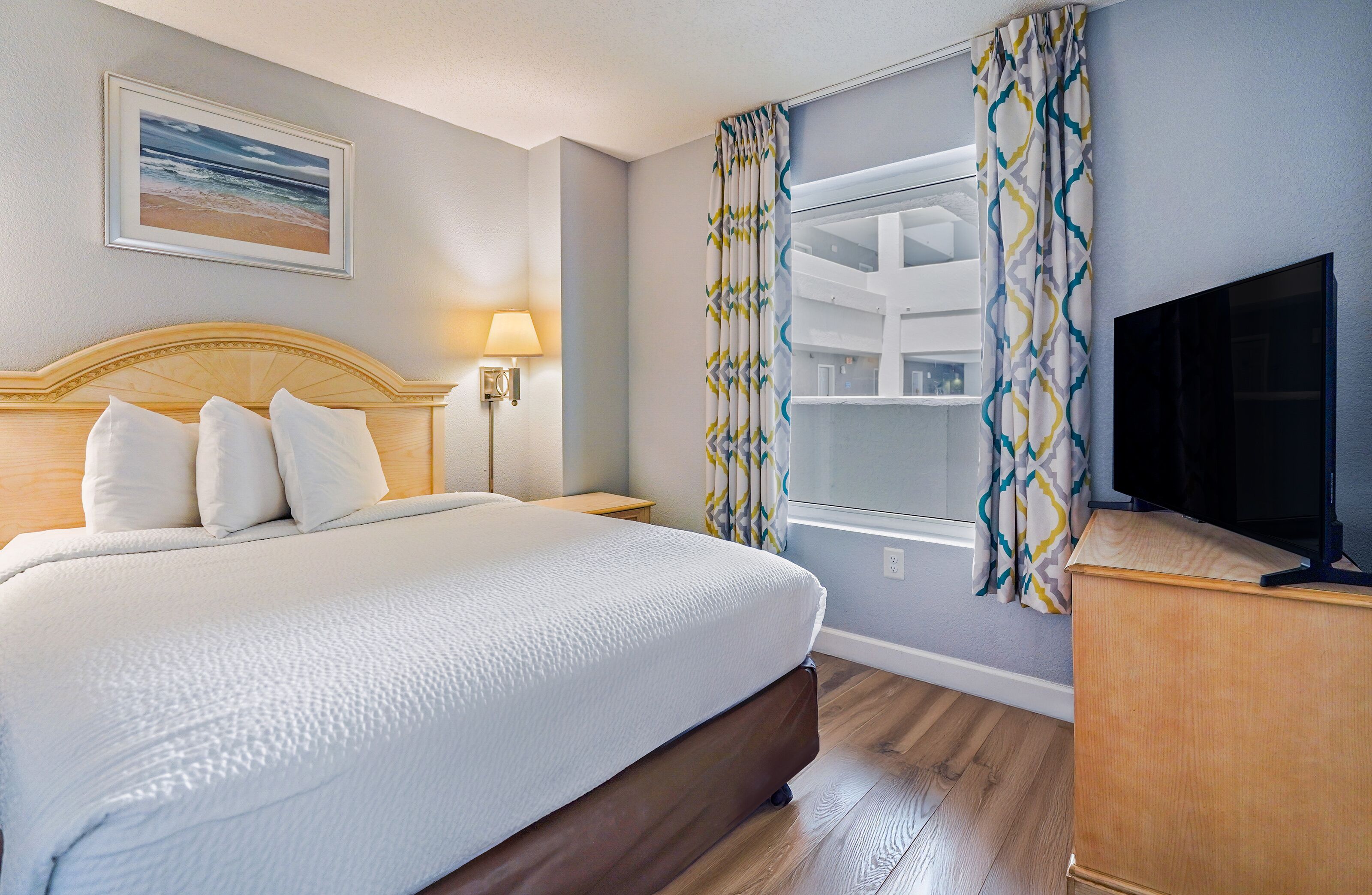 A hotel room with a large bed, a TV, a lamp, and a window with patterned curtains.