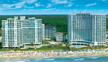 A beachfront hotel complex with multiple high-rise buildings, a pool area, and a sandy beach with umbrellas.