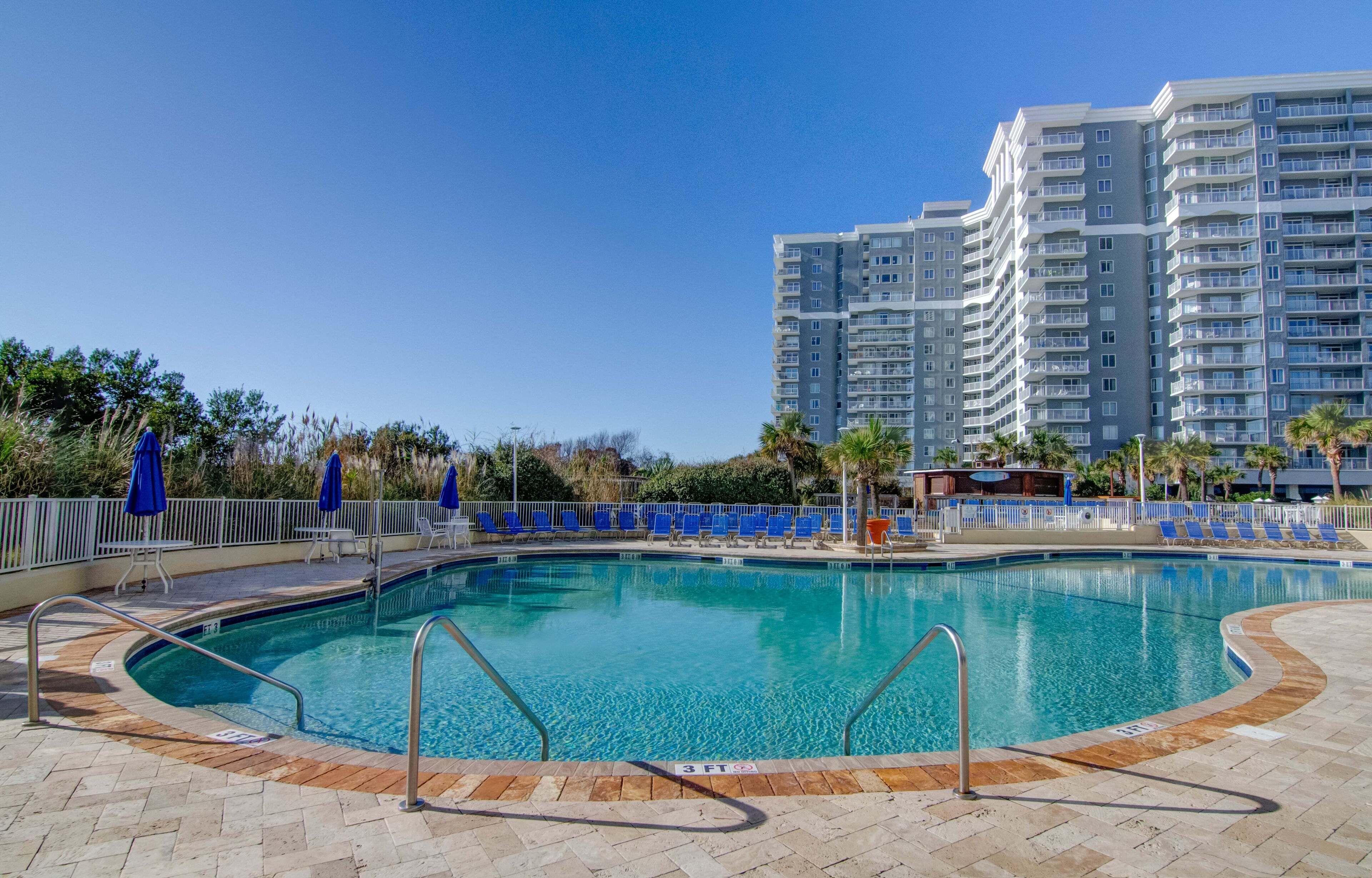 A swimming pool with a 3 FT depth marker, surrounded by blue chairs and umbrellas, with a high-rise building in the background.