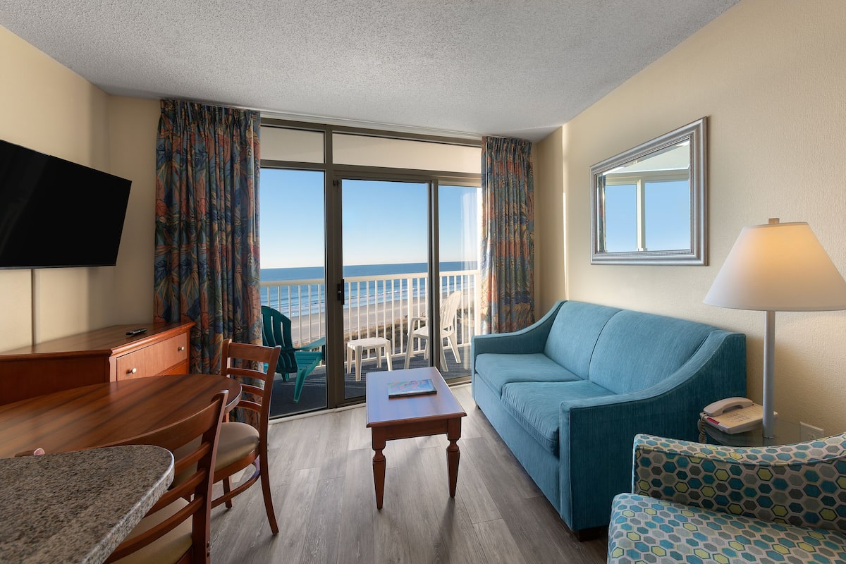 A hotel room with a balcony, a flat-screen TV, a sofa, a coffee table, and a lamp.
