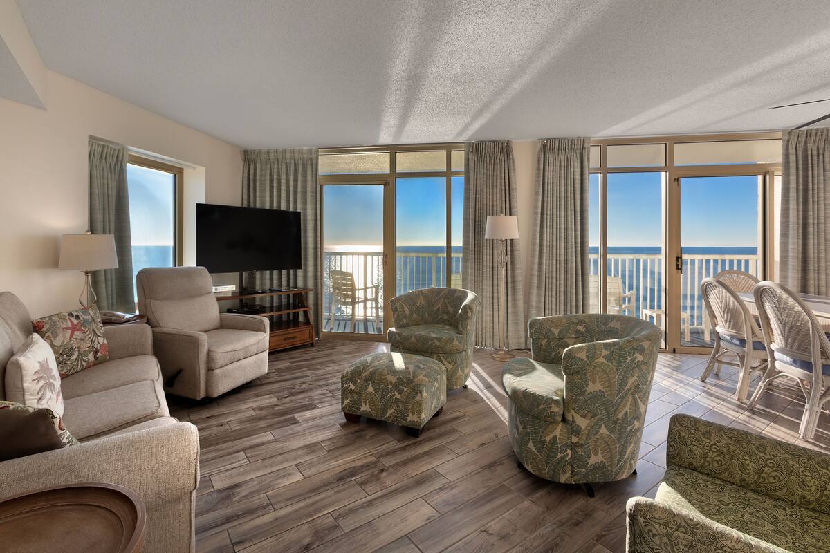 A spacious living room with a large flat-screen TV, comfortable seating, and a balcony with a view of the sea.