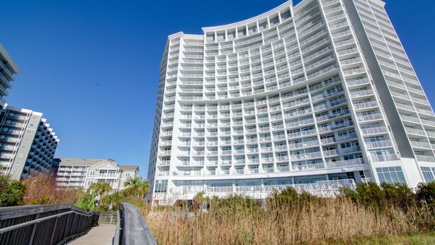 A modern multi-story hotel with a curved facade, balconies, and a wooden boardwalk in the foreground. - Sea Watch Resort (Myrtle Beach)