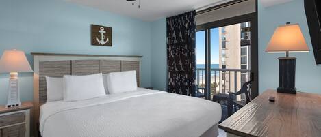 A hotel room with a bed, two bedside tables with lamps, a ceiling fan, a window with a view of buildings, and a wooden table.