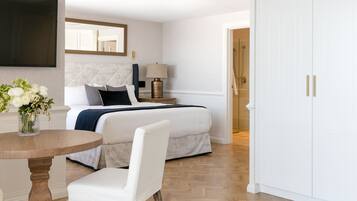 Une chambre d’hôtel comprenant un lit, une table de chevet avec une lampe, une chaise, une table ronde en bois ornée d’un vase de fleurs et un téléviseur fixé au mur.