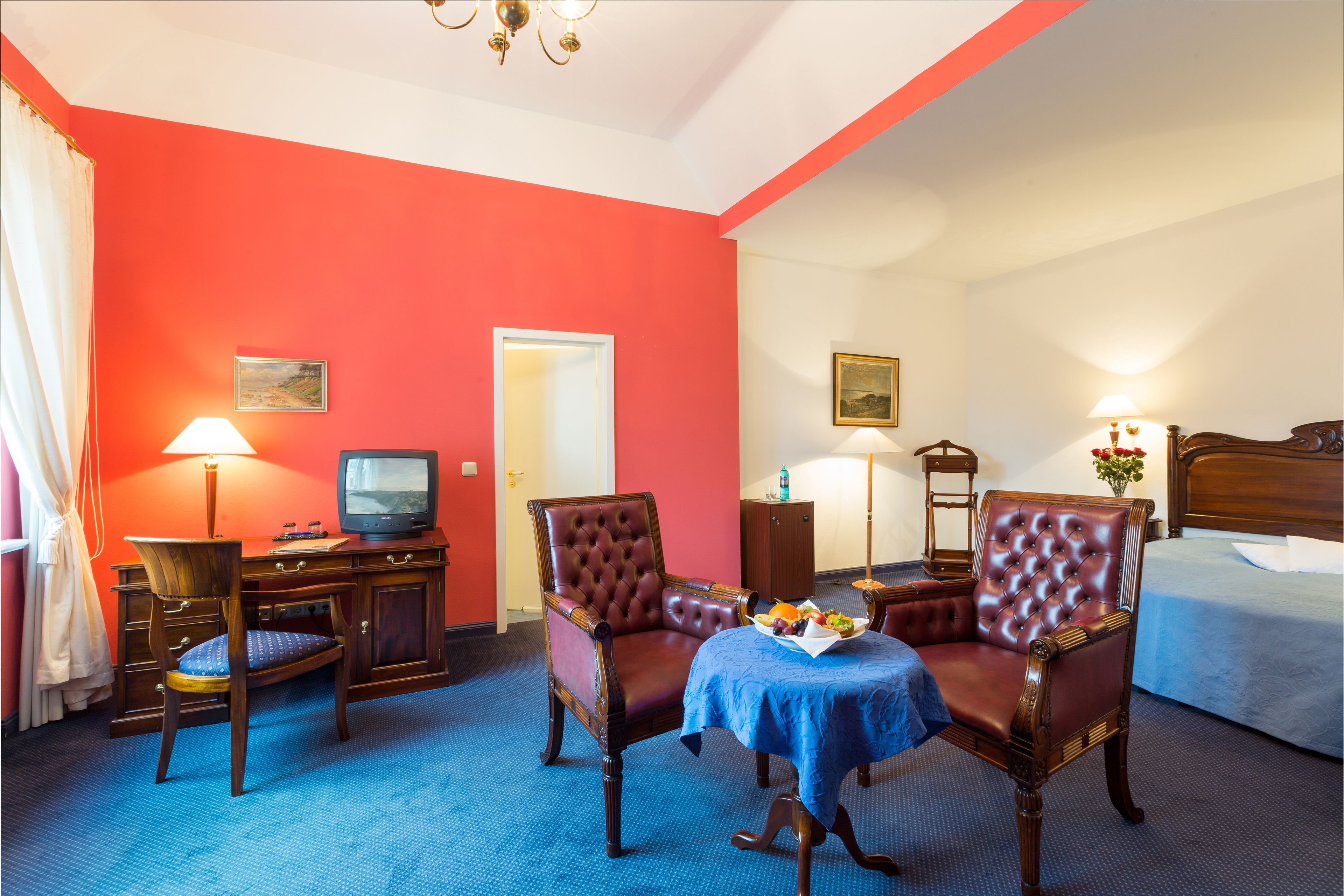 A hotel room with a red wall, a bed, a desk with a television, two leather chairs, a small table with fruit, and a blue carpet.