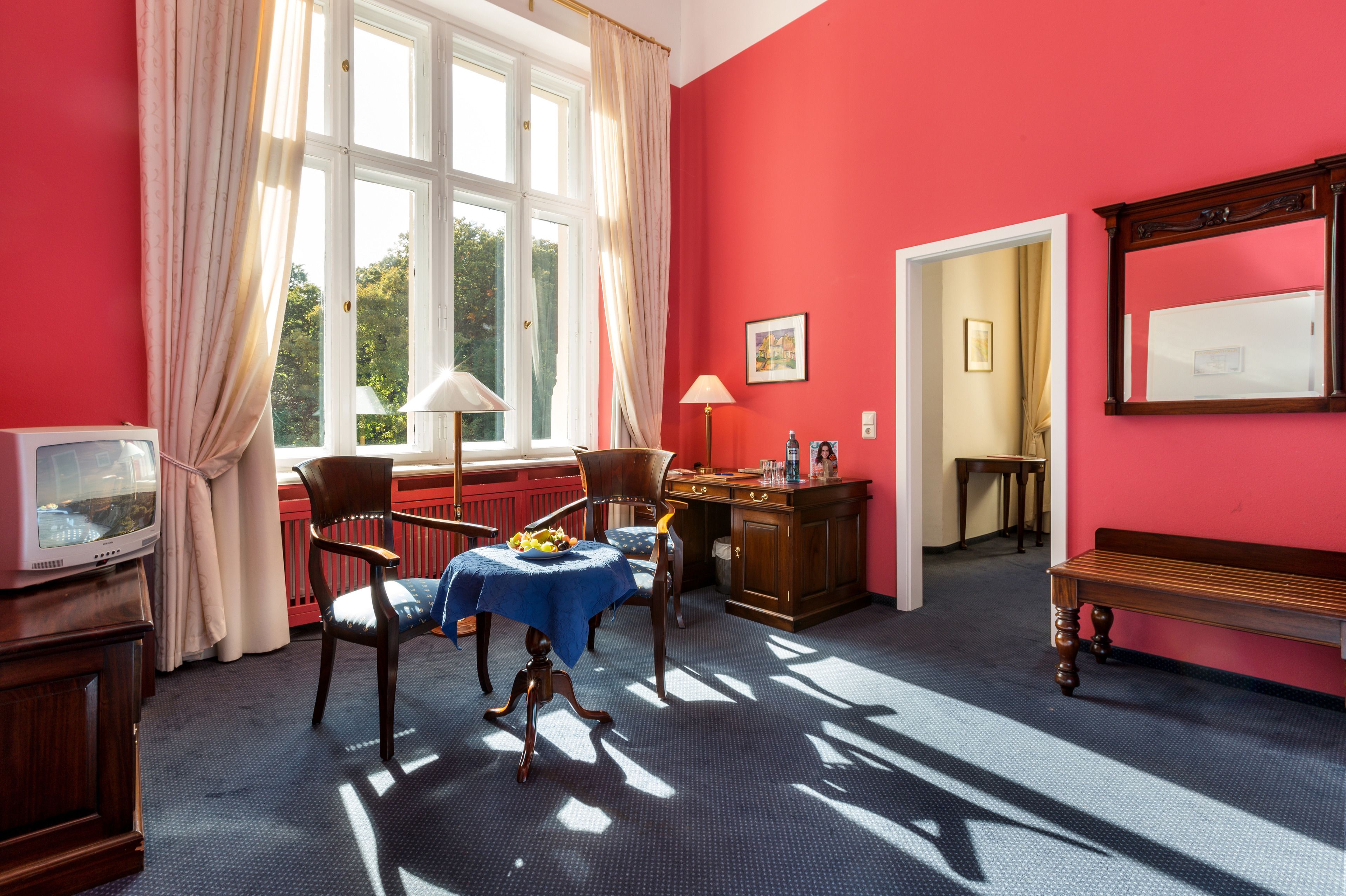 Una habitación con una pared roja, un escritorio de madera, una silla, una mesa con mantel azul, una lámpara, un televisor y un banco.