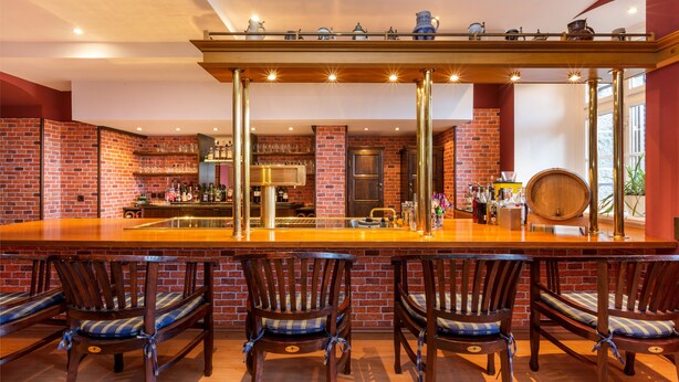 A bar area with a long wooden counter, four wooden bar stools with blue cushions, and a brick wall backdrop. - Schlosshotel Ralswiek (Ralswiek)